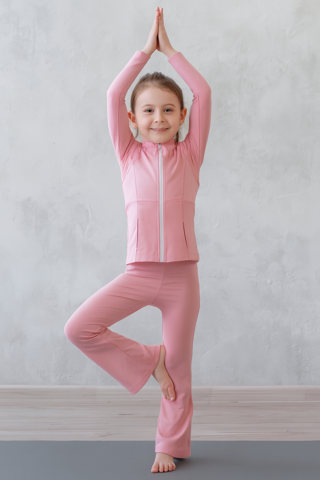 Child in pink outfit standing on one leg with hands raised against a light gray background